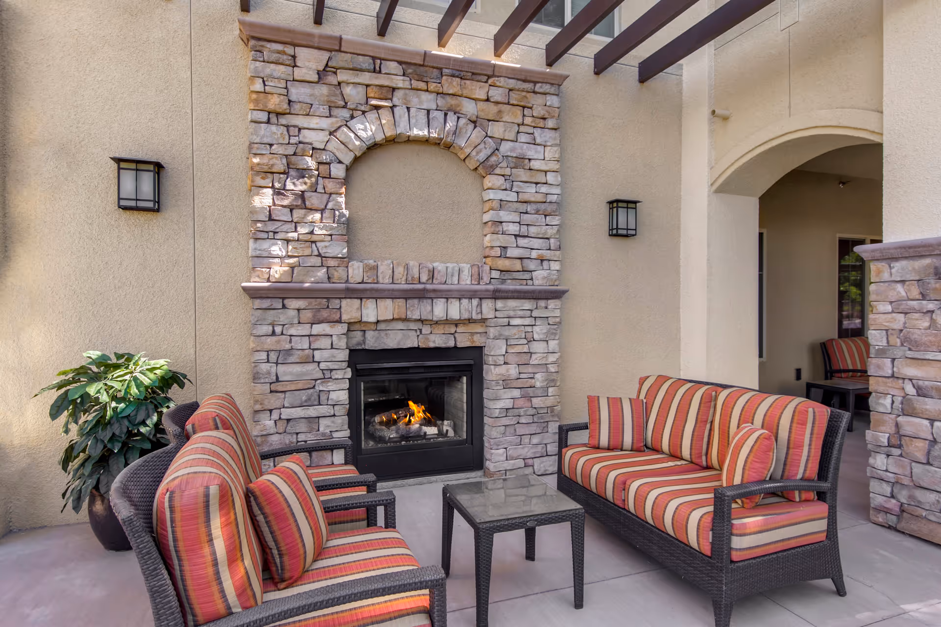 Outdoor seating area with two cushioned wicker sofas and a small glass-top table in front of a stone fireplace with a fire burning. The area is covered by a pergola and has beige walls with two wall-mounted lights and a potted plant on the left side.