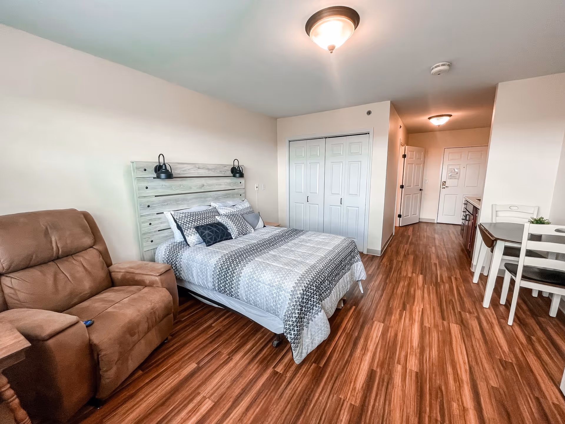 A senior living facility room featuring a bed with patterned bedding, a brown recliner chair, a small dining table with two chairs, and a kitchenette area. The room has wood flooring, white walls, and ceiling lights. There is a closet with white sliding doors and an entrance door visible in the background.