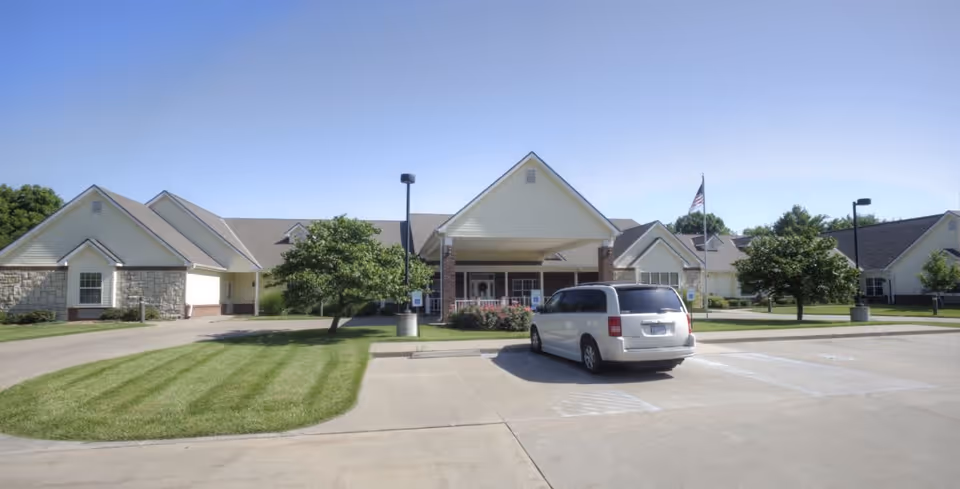 Front exterior view of a senior living facility named Vintage Park at Osawatomie, showing a single-story building with a covered entrance, a white van parked in front, green lawns, trees, and a clear blue sky.