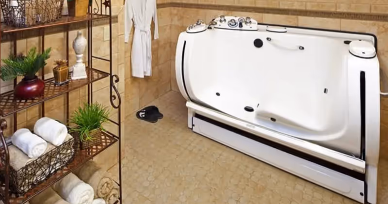 Accessible walk-in bathtub in a tiled bathroom with shelving holding rolled towels and potted plants.