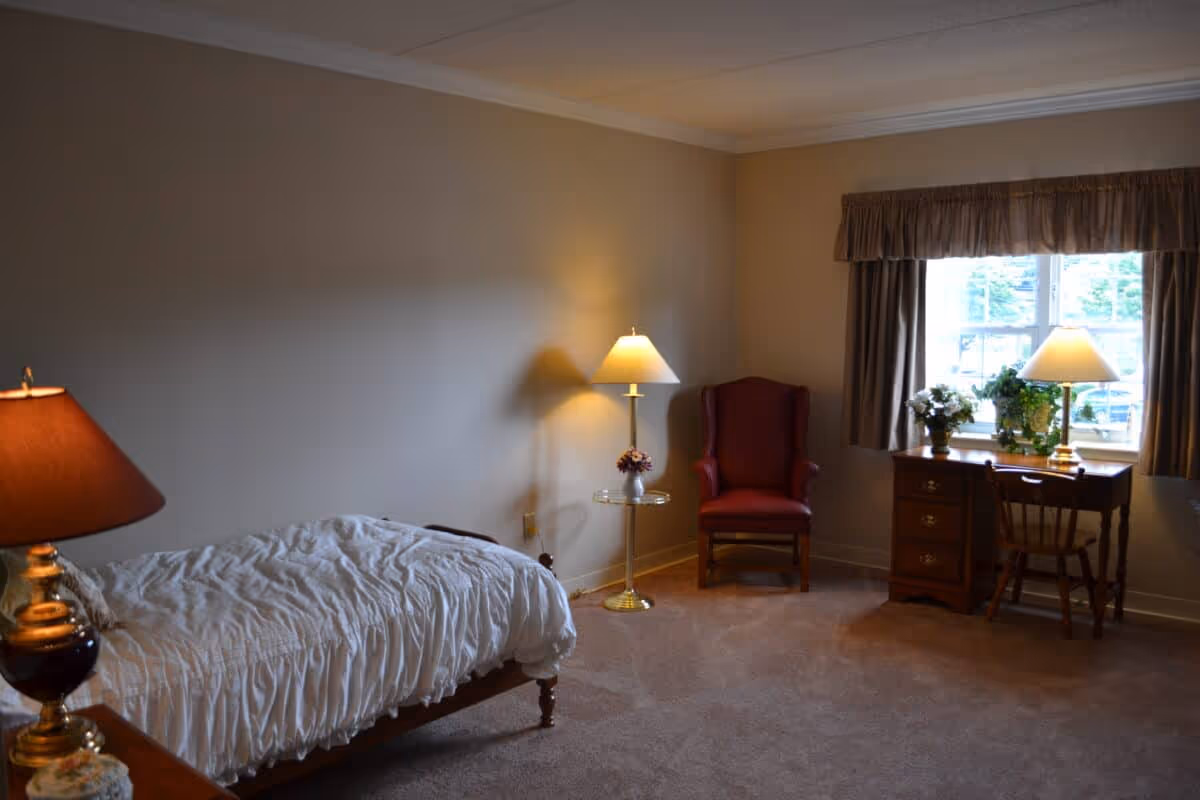 A senior living bedroom with a single bed covered in a white comforter, a wooden nightstand with a lamp, a red armchair, a small round side table with a flower vase, a wooden desk with a chair, and a window with curtains letting in natural light. The room has beige walls and carpeted floor.