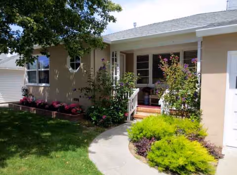 Single-story beige senior living facility with front porch, walkway, and flowering shrubs.