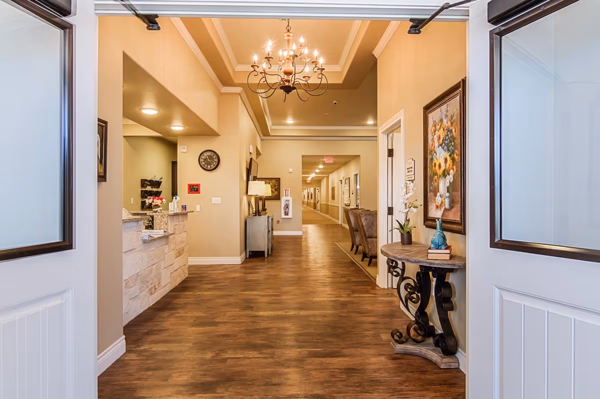 View through double doors into a well-lit hallway of an assisted living facility with wood flooring, beige walls, a chandelier, a reception desk on the left, and a decorative table with a flower vase and framed painting on the right.