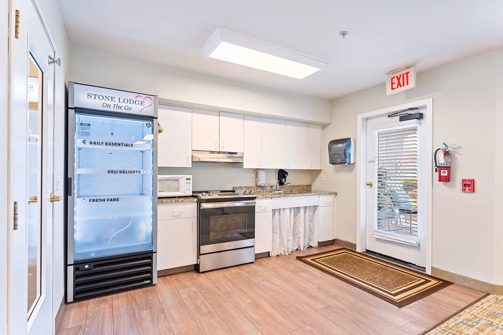 A clean and bright kitchen area with white cabinets, a stainless steel stove, microwave, and a granite countertop. To the left, there is a refrigerated display case labeled 'STONE LODGE On The Go' with sections for Daily Essentials, Deli Delights, and Fresh Fare. A door with blinds and an exit sign is visible on the right side, along with a fire extinguisher and fire alarm on the wall. The floor is wood-patterned, and there is a brown and beige rug near the door.