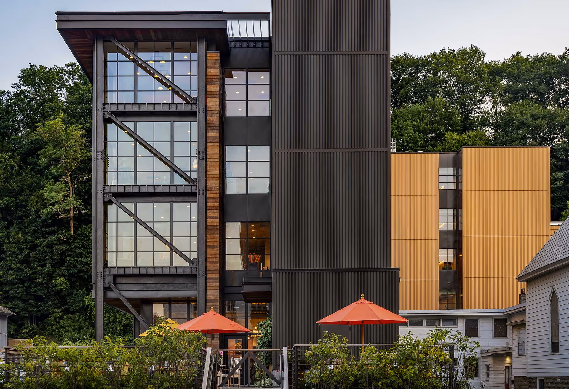 Exterior view of a modern multi-story building with large glass windows and metal framework, surrounded by greenery. In front of the building are two red patio umbrellas and some plants.