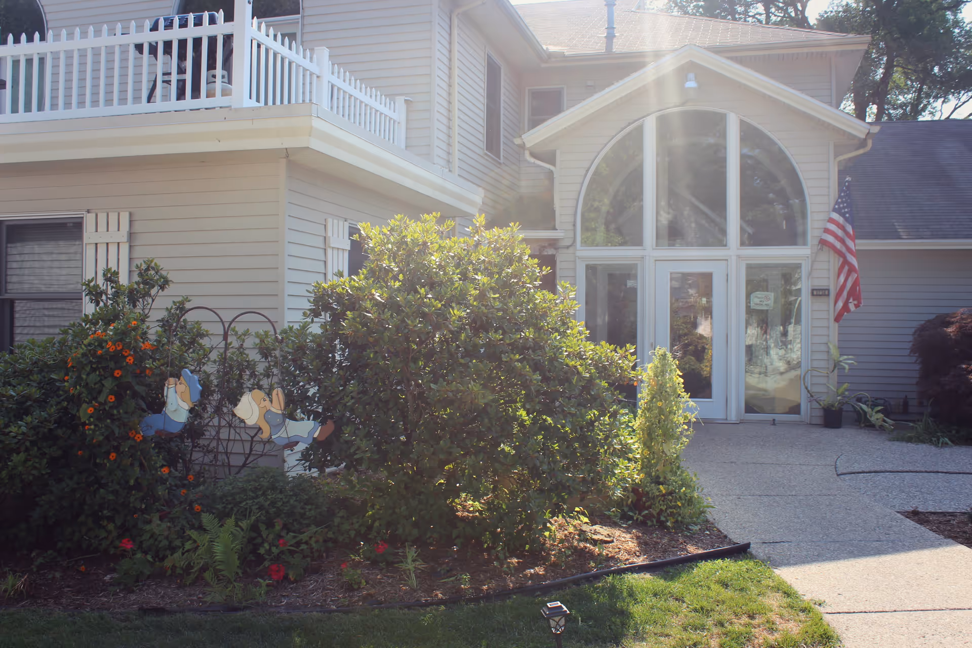 Front entrance of a light-colored building with large arched windows, an American flag, and landscaping in the foreground.