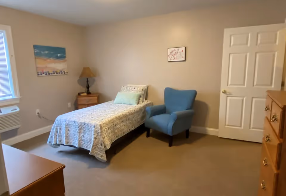 A simple bedroom with a single bed covered with a patterned quilt, a blue upholstered armchair, a wooden nightstand with a lamp, a wooden dresser, and two framed pictures on beige walls. There is a window with blinds and an air conditioning unit below it.