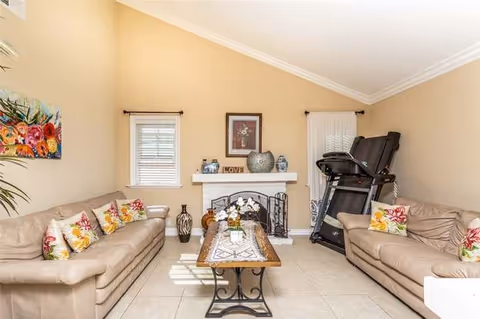 A cozy living room with two beige leather sofas facing each other, each adorned with colorful floral cushions. A wooden coffee table with a decorative runner and a small flower arrangement sits in the center on a tiled floor. The room features a white fireplace with decorative items on the mantel, two small windows with blinds, and a treadmill positioned against the right wall. The walls are painted a warm beige color, and there is a colorful floral painting on the left wall.