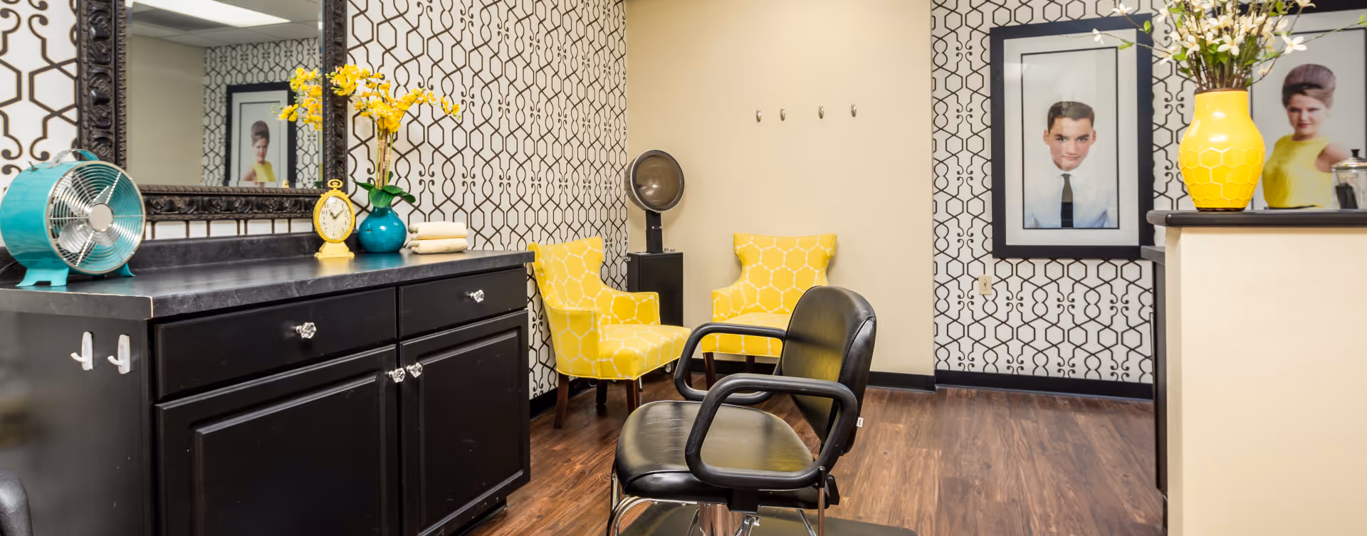 Stylish interior salon area with black styling chairs, a large mirror over a black cabinet, patterned wallpaper, yellow accent chairs and vases, and framed portraits on the wall.