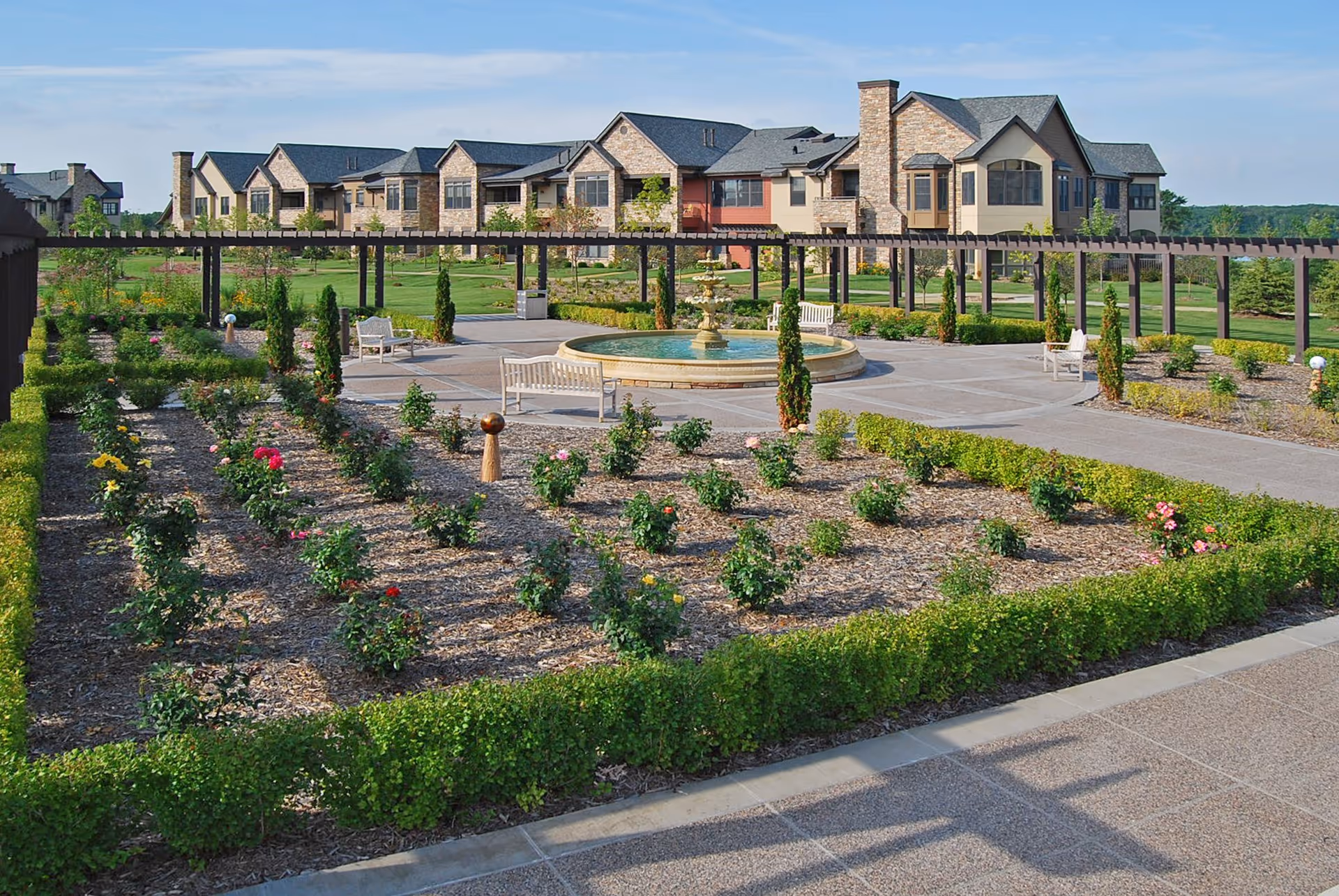 A landscaped garden area with neatly arranged flower beds, a central circular fountain, white benches, and a pergola structure. In the background, there is a large building with stone and beige exterior walls under a clear blue sky.