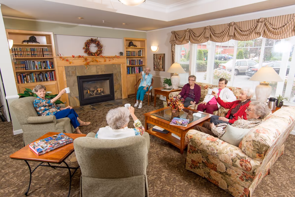 A group of elderly women sitting in a cozy living room with a fireplace, bookshelves, and large windows. They are seated on armchairs and floral-patterned sofas, holding mugs and smiling, enjoying a social gathering.