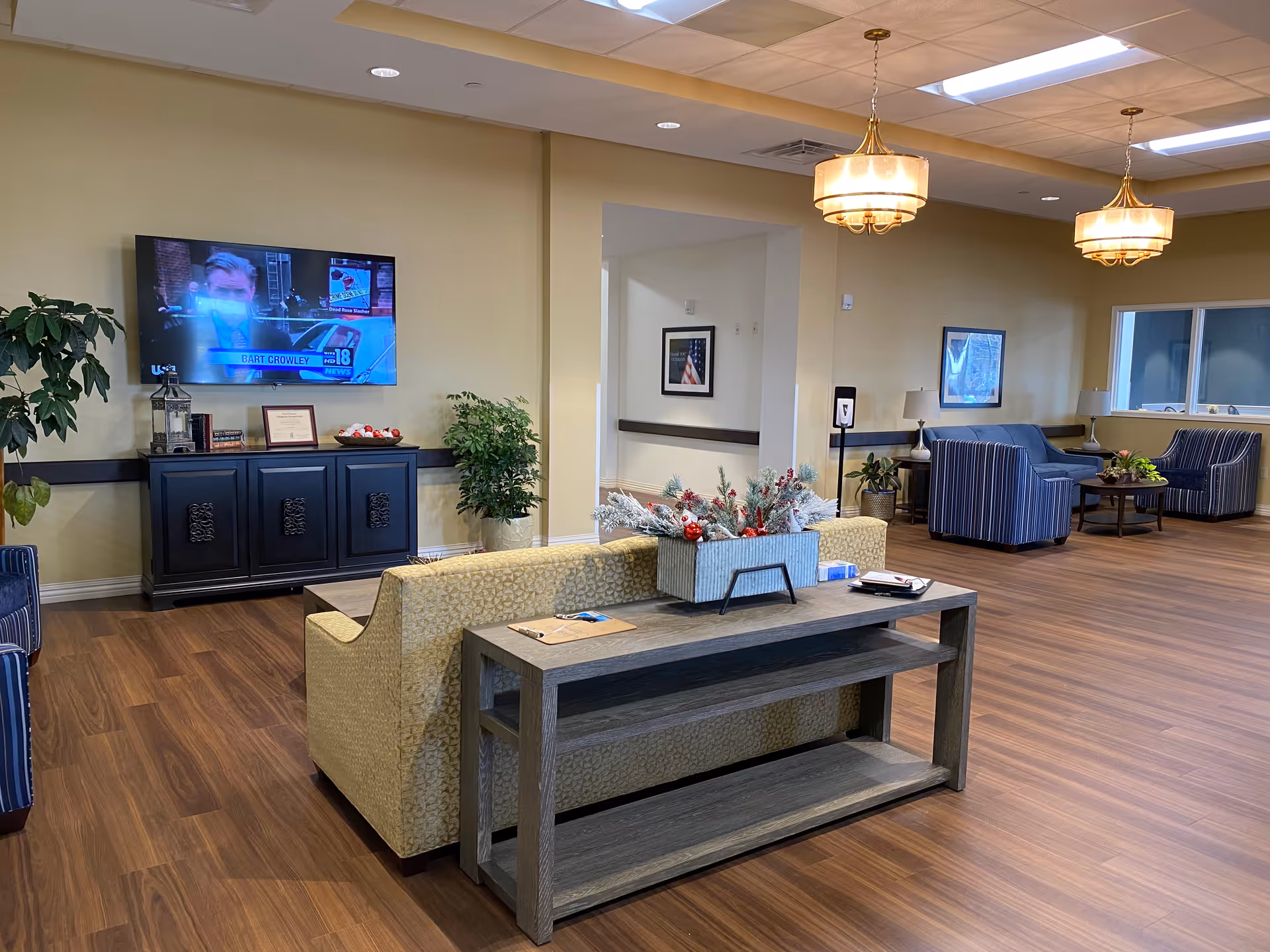A spacious and well-lit nursing home common area with wooden flooring, a mounted flat-screen TV showing a news broadcast, a black cabinet with decorative items, a beige patterned sofa with a wooden console table behind it, and two blue striped armchairs with a small round table. The room features warm yellow walls, ceiling lights, and some potted plants.