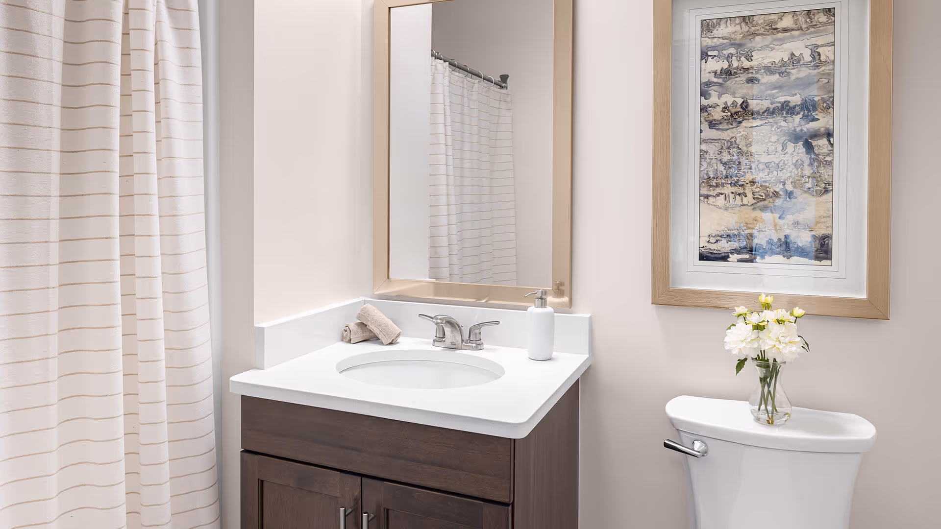 A clean and modern bathroom featuring a white countertop with an oval sink, a silver faucet, a soap dispenser, and a rolled towel. There is a large mirror above the sink, a shower curtain with subtle stripes to the left, and a white toilet with a vase of white flowers on top. A framed abstract artwork hangs on the wall above the toilet.