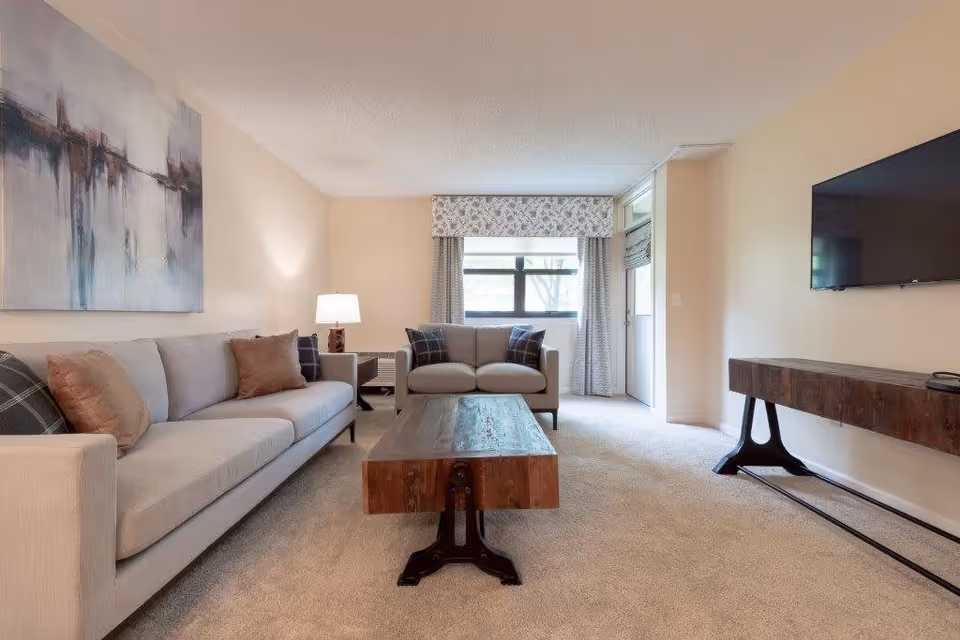 A bright living room with a beige carpet, a long light gray sofa with brown and plaid pillows, a matching loveseat with plaid pillows, a wooden coffee table with black metal legs, a wooden console table, a wall-mounted flat screen TV, a window with patterned curtains, and a door leading outside.