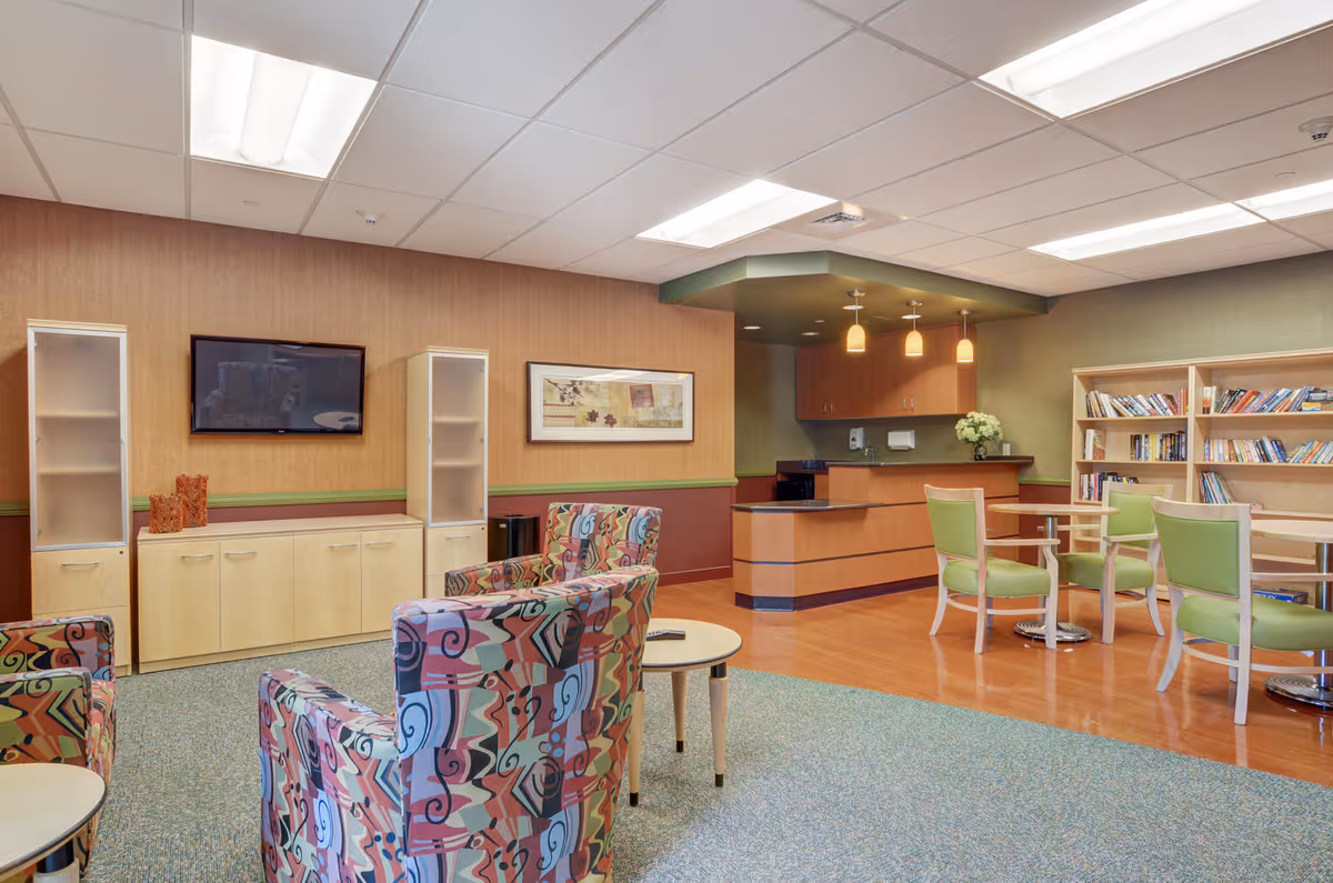 A cozy common area in a senior living facility featuring colorful patterned armchairs around small round tables, a wall-mounted flat screen TV, light wood cabinetry with glass-fronted shelves, a small kitchenette with hanging pendant lights, and bookshelves filled with books.