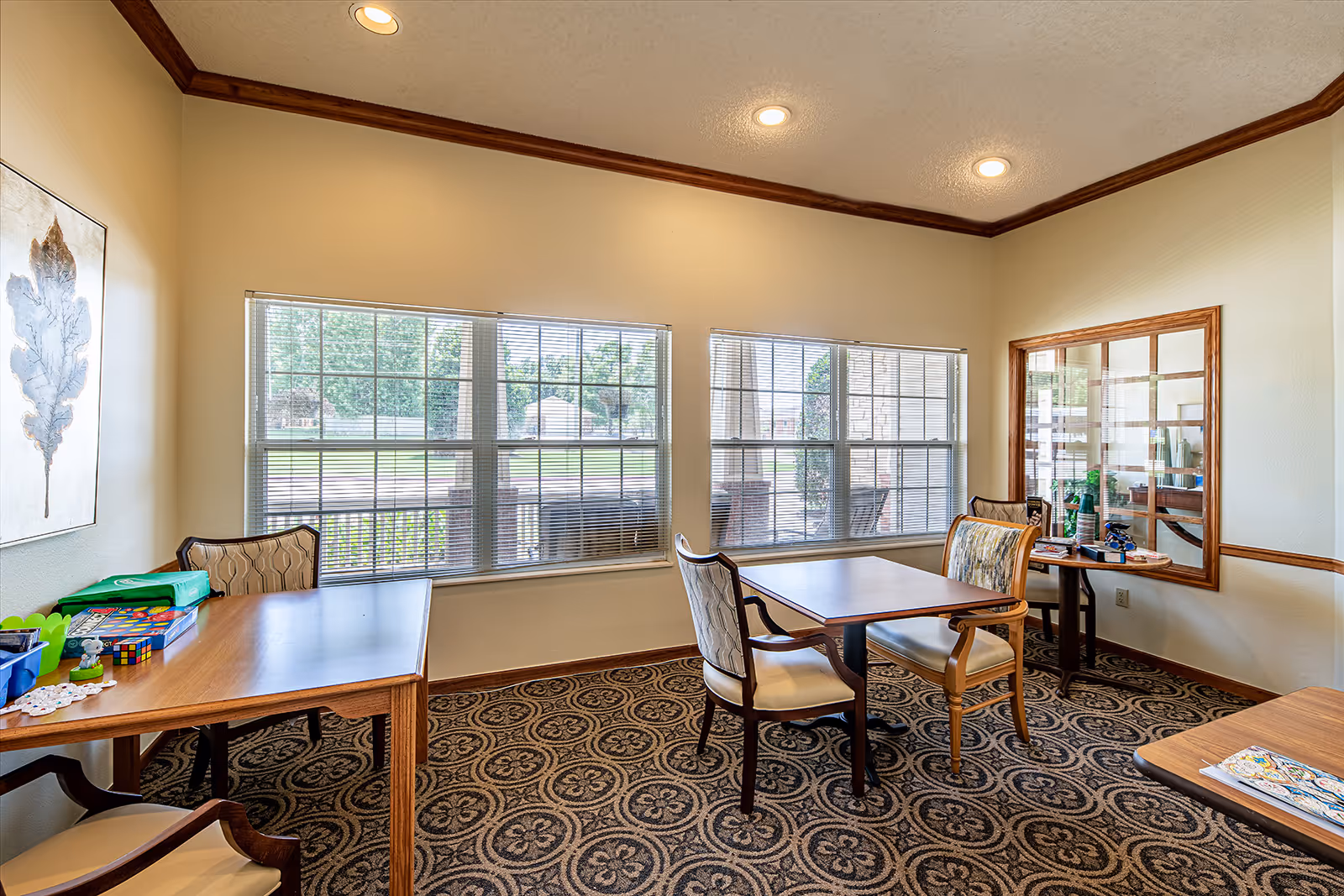 A well-lit room with large windows featuring horizontal blinds, several wooden tables with cushioned chairs, a patterned carpet, and a framed artwork of a leaf on the wall. The room has beige walls with wooden trim and recessed ceiling lights.