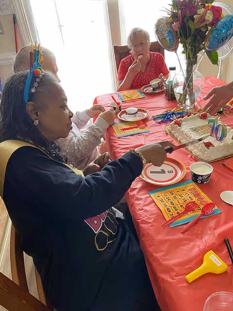 Three elderly women sitting around a table covered with a red tablecloth, celebrating a birthday. One woman wearing a birthday crown is cutting a cake decorated with a '70' candle. There are bingo cards, plates, utensils, and a vase with flowers and balloons on the table. One woman is eating a cupcake.