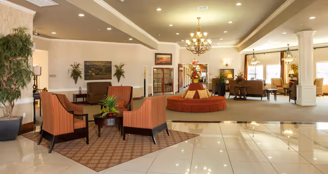 A spacious and well-lit living room area in a senior living facility featuring multiple seating arrangements including striped armchairs around a small round table on a patterned rug, a circular cushioned bench with decorative pillows in the center, and additional sofas and chairs near large windows. The room has polished tile flooring, beige walls with white wainscoting, decorative plants, framed artwork, and a chandelier hanging from the ceiling.