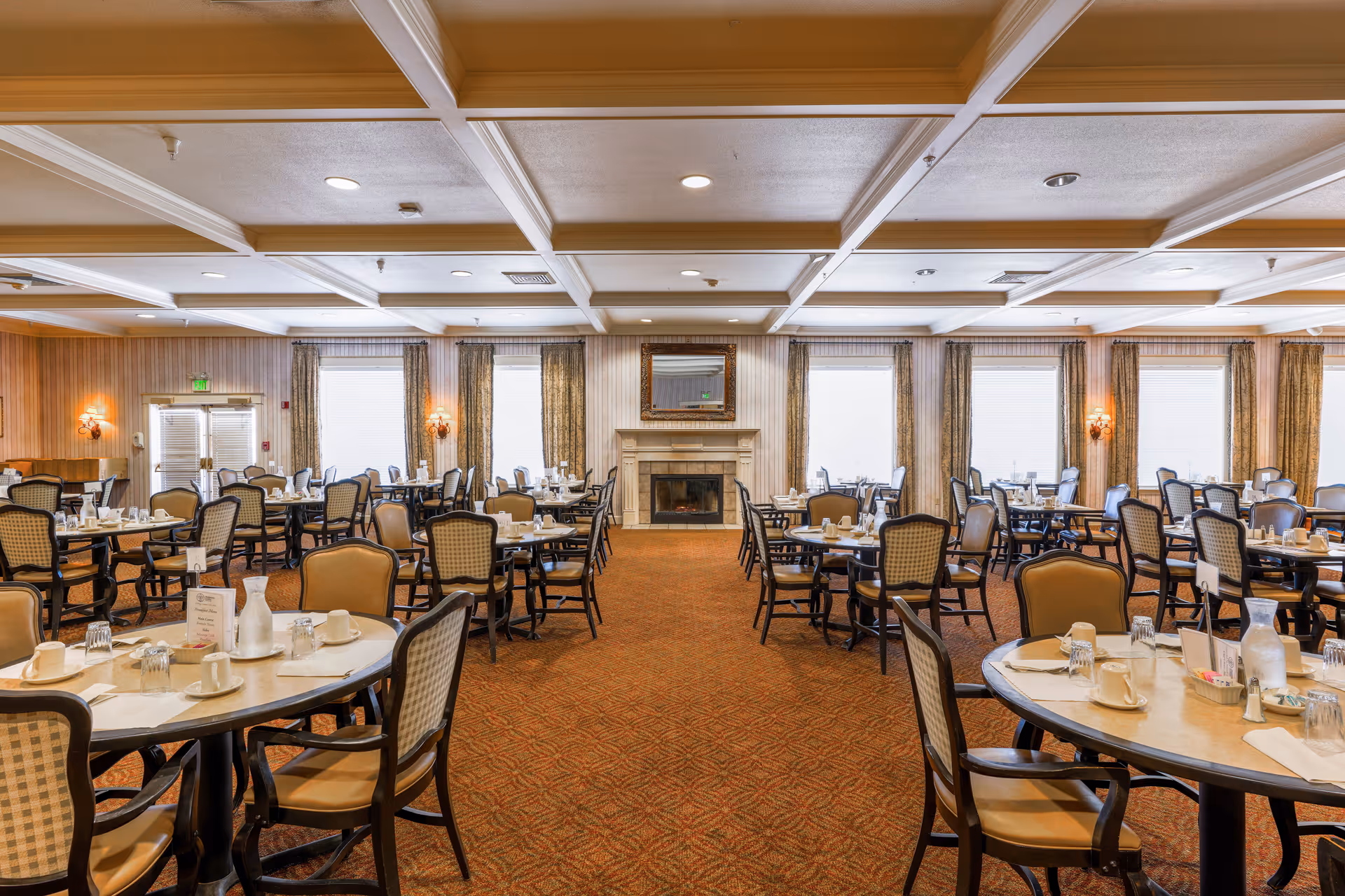 A spacious dining room with multiple round tables and chairs arranged neatly. Each table is set with cups, glasses, napkins, and condiments. The room features a coffered ceiling, large windows with curtains, wall sconces, and a central fireplace with a mirror above it. The carpet has a patterned design in warm tones.