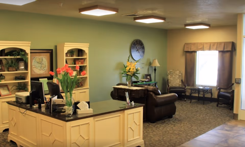 Reception area with a cream-colored desk featuring a vase of red tulips, two computer monitors, and office supplies. Behind the desk are two cream-colored bookshelves with decorative items and plants. To the right, there is a seating area with a brown leather couch, two armchairs, a side table with a lamp, and a large window with brown curtains. The walls are painted green and beige, and the floor is carpeted with a patterned design.