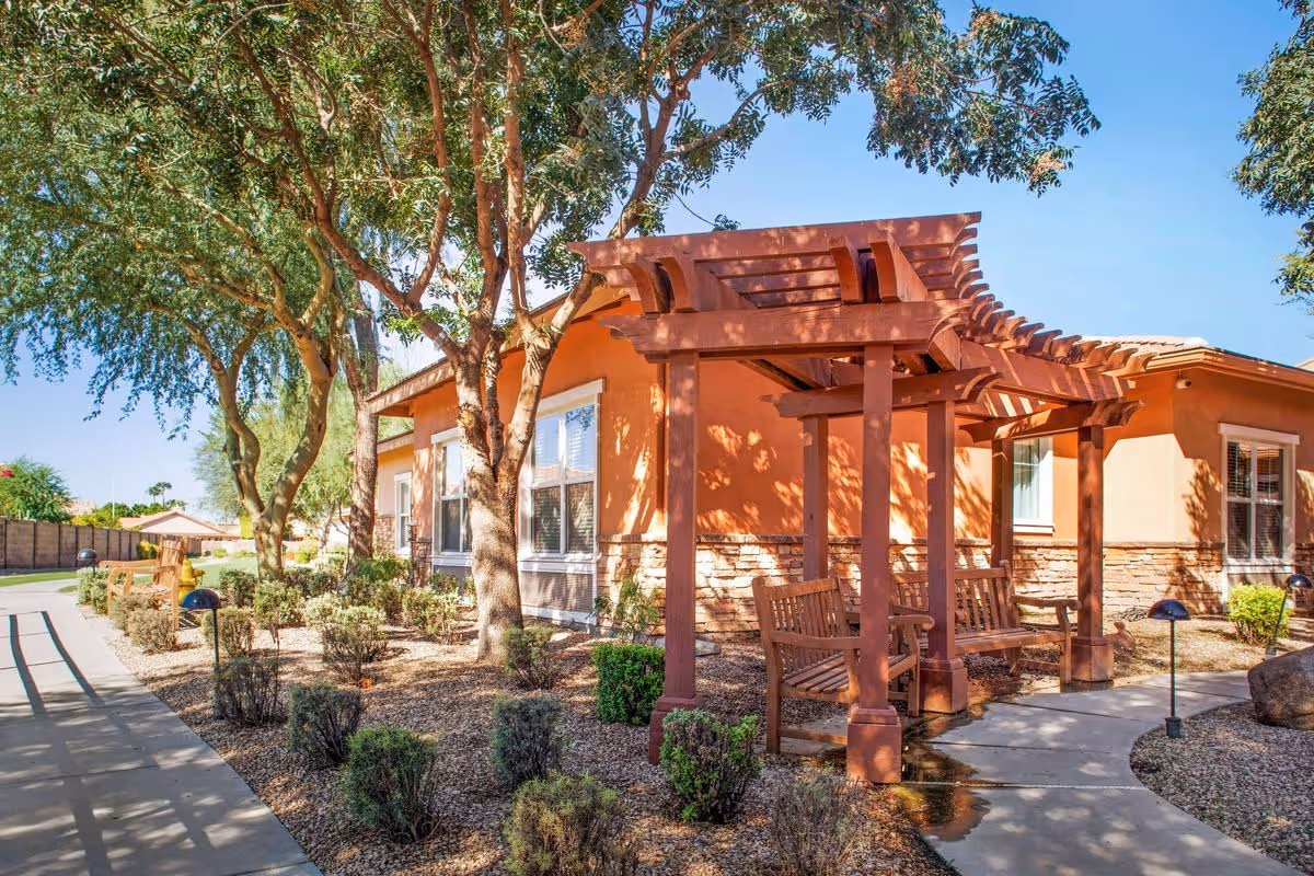 Wooden pergola with benches beside a landscaped walkway and stucco building under trees.