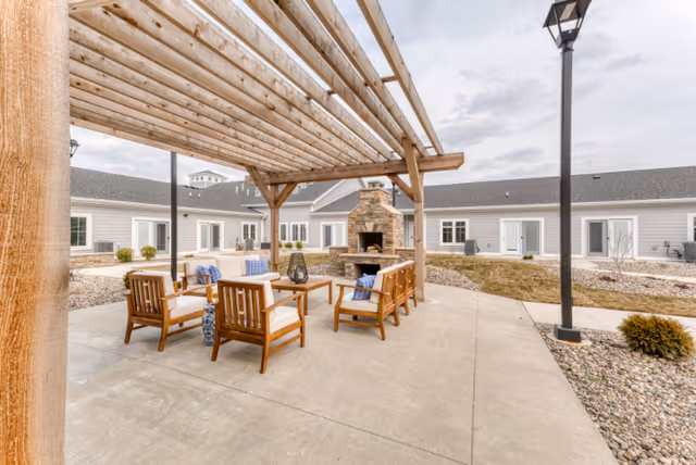 Outdoor seating area at Cedarhurst Senior Living of Fort Wayne featuring wooden chairs with cushions arranged around a coffee table under a wooden pergola, with a stone fireplace in the background and a single lamp post on the right side. The area is surrounded by a building with multiple windows and doors, and landscaping with rocks and small bushes.