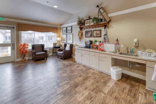 A bright room with wood flooring featuring two brown armchairs near a large window with a checkered valance. To the right, there is a long countertop with cabinets underneath, a sink, a soap dispenser, and various decorative items including plants and framed pictures on the wall above. A small trash can is placed under the sink. The room has a door leading outside and a small table with a lamp between the chairs.