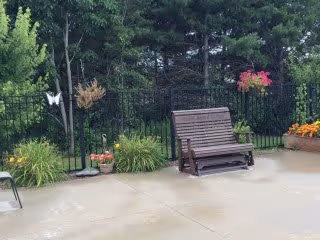 Outdoor patio area with a wooden bench, potted plants, hanging flower baskets, and greenery including trees and bushes in the background.
