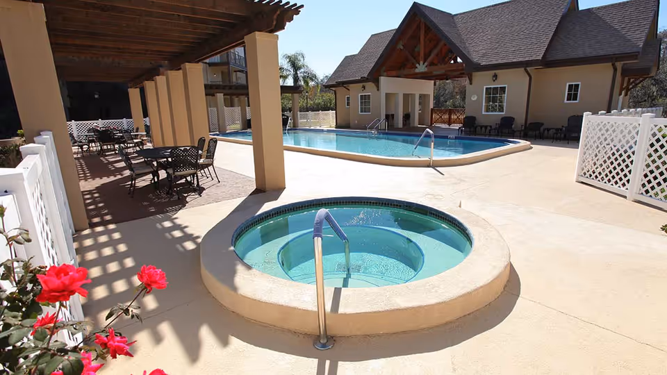 Outdoor pool deck featuring a round hot tub in the foreground, a larger swimming pool, a pergola with tables and chairs, and a building in the background.