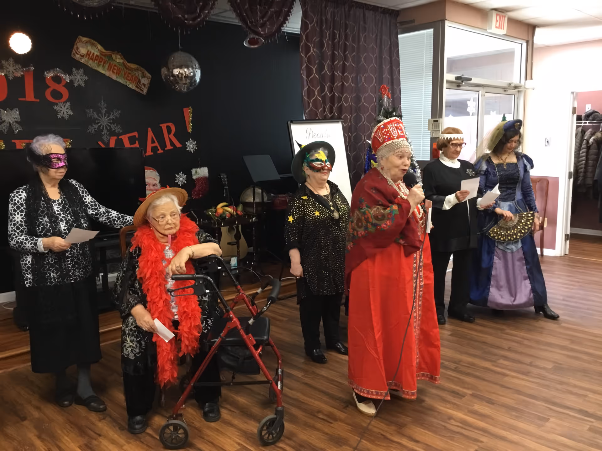 A group of elderly women dressed in festive costumes and masks are standing in a room with wooden flooring. One woman in a red traditional outfit is speaking into a microphone. The background has New Year decorations including snowflakes and a sign that says 'Happy New Year 2018'.