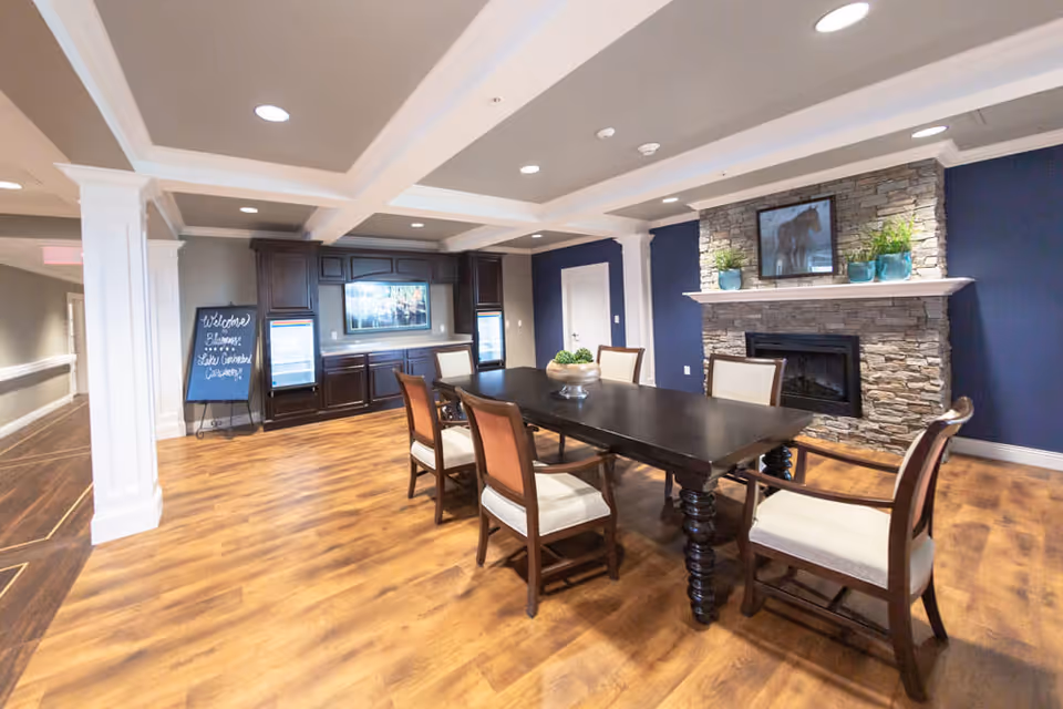 A spacious dining room with a long dark wooden table surrounded by six cushioned chairs. The room features a stone fireplace with a framed picture and potted plants on the mantel. The walls are painted navy blue and the floor has a wood finish. There is a cabinet with a TV and two refrigerators in the background, and a chalkboard sign near the entrance.