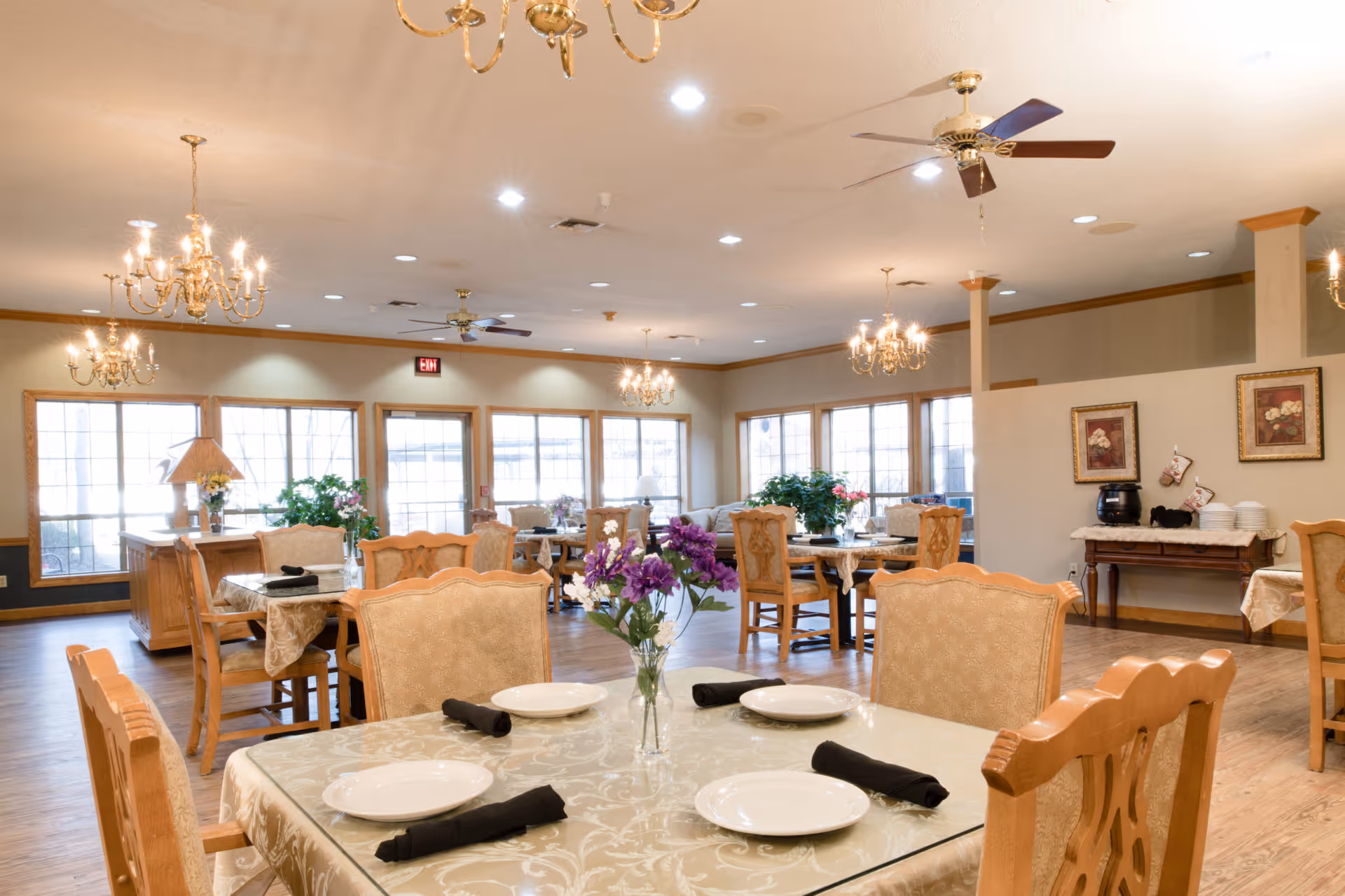A bright and spacious dining room in a senior living facility with multiple tables covered in patterned tablecloths, each set with white plates and black napkins. The room features wooden chairs, chandeliers, ceiling fans, large windows letting in natural light, and decorative plants and framed artwork on the walls.