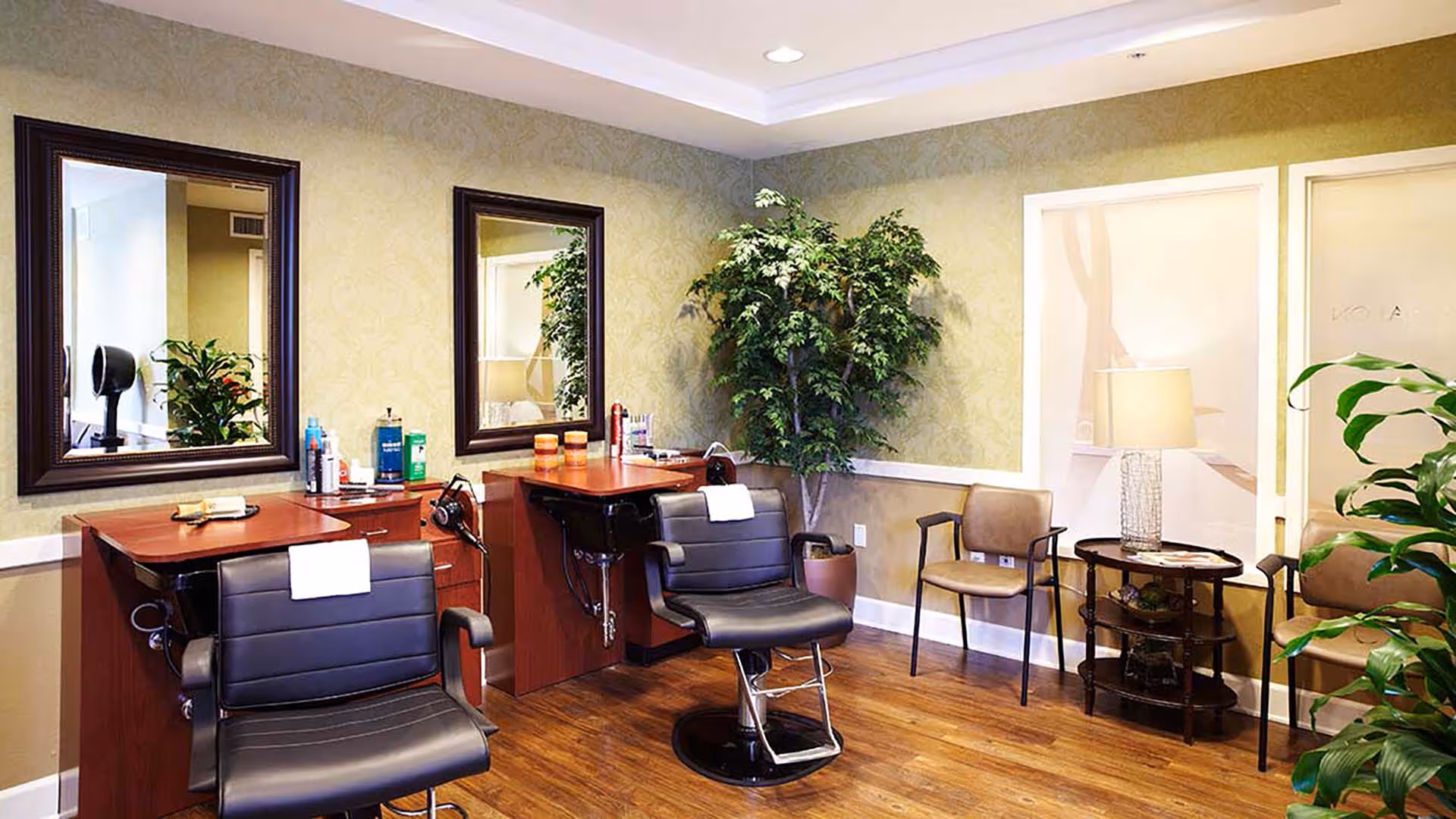 Interior view of a salon area with two black salon chairs in front of wooden counters with mirrors. Various hair care products are placed on the counters. There are additional chairs and a small table with a lamp in the corner, along with potted plants and light green wallpaper.