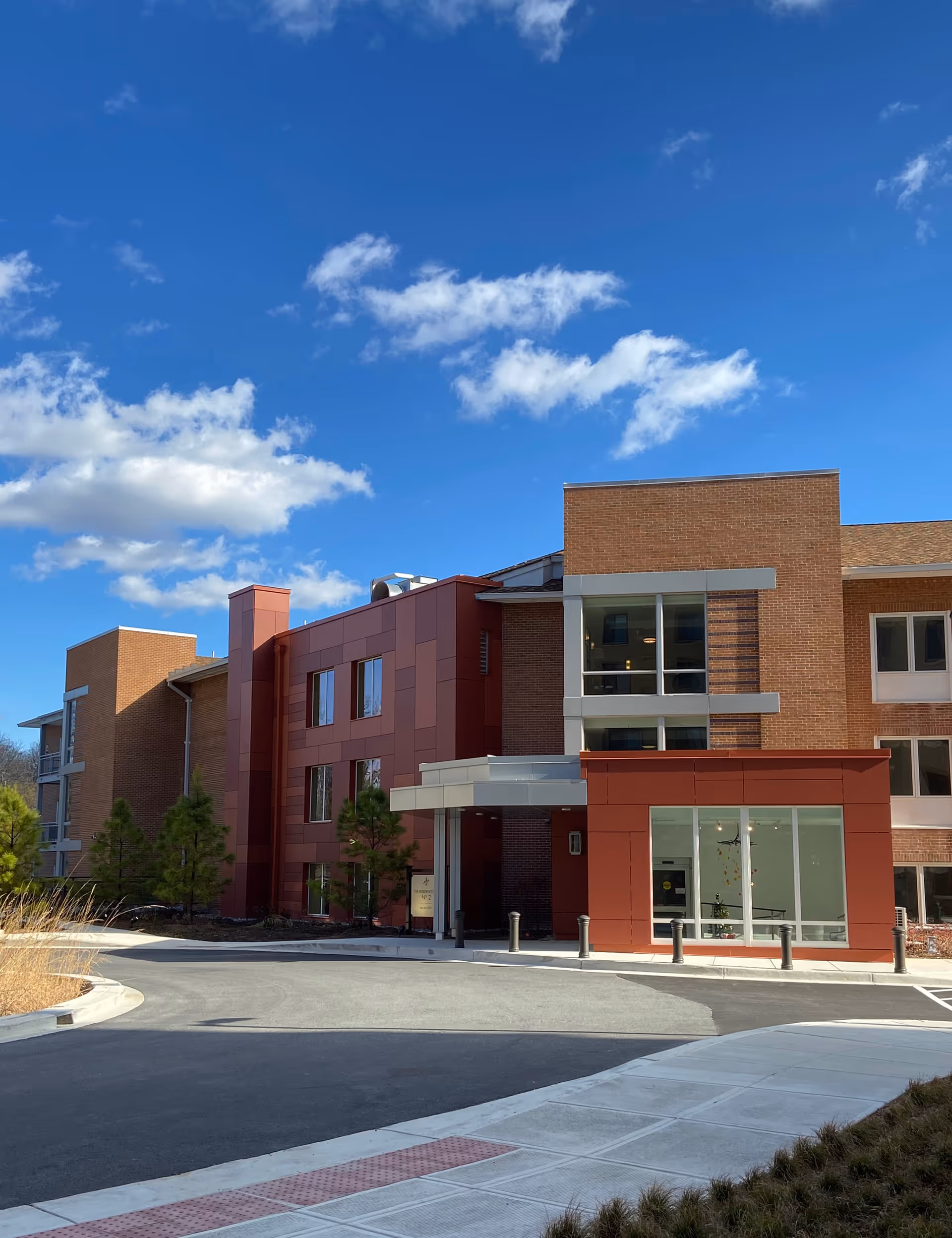 Entrance of a multi-story brick and red-paneled senior living building with a circular driveway under a blue sky.