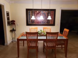 Dining area with a wooden table and six matching chairs, three pendant lights hanging above the table, a decorative centerpiece on the table, a large framed artwork on the wall, and a small plant stand with flowers in the corner.