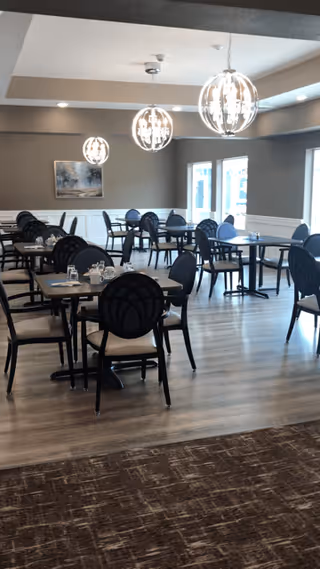 A dining room with multiple tables and black chairs arranged neatly. The room has modern spherical pendant lights hanging from the ceiling, large windows letting in natural light, and a painting on the far wall. The floor is wooden with a carpeted area in the foreground.