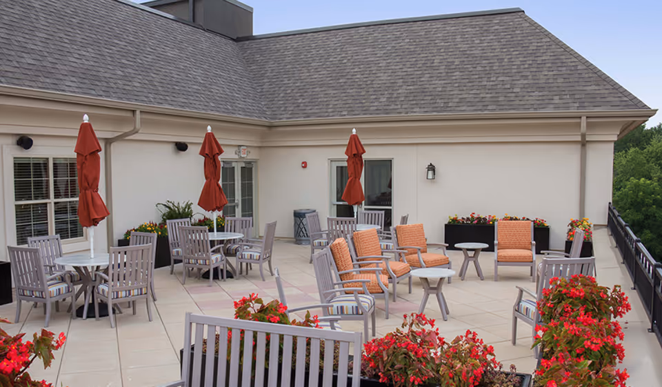 Rooftop terrace with multiple tables and chairs, folded red umbrellas, and planters of red flowers.