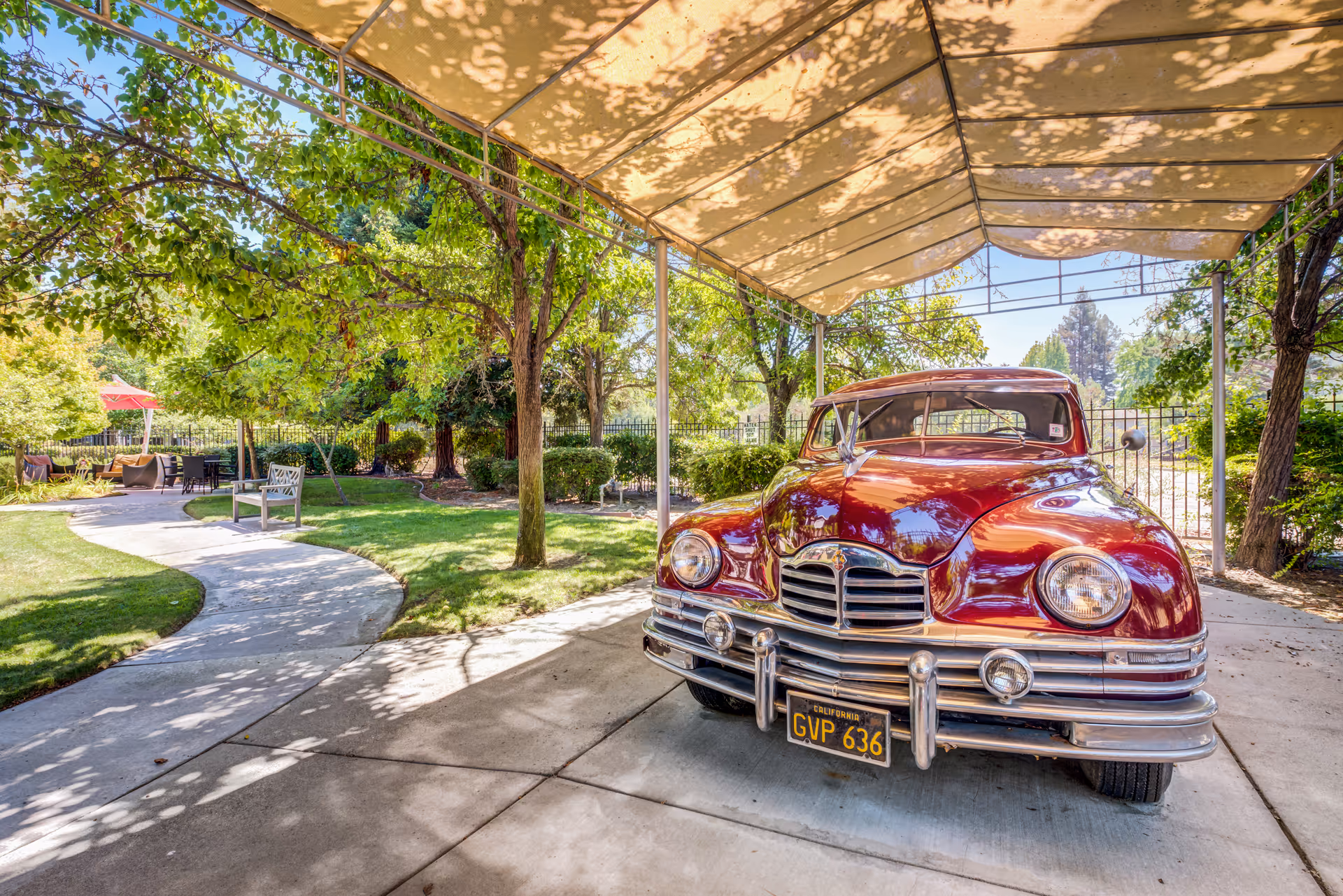 A vintage red car with a California license plate parked under a beige canopy in a shaded outdoor area. Surrounding the car are green trees, bushes, and a curved concrete pathway leading to outdoor seating with chairs and a red umbrella in the background.