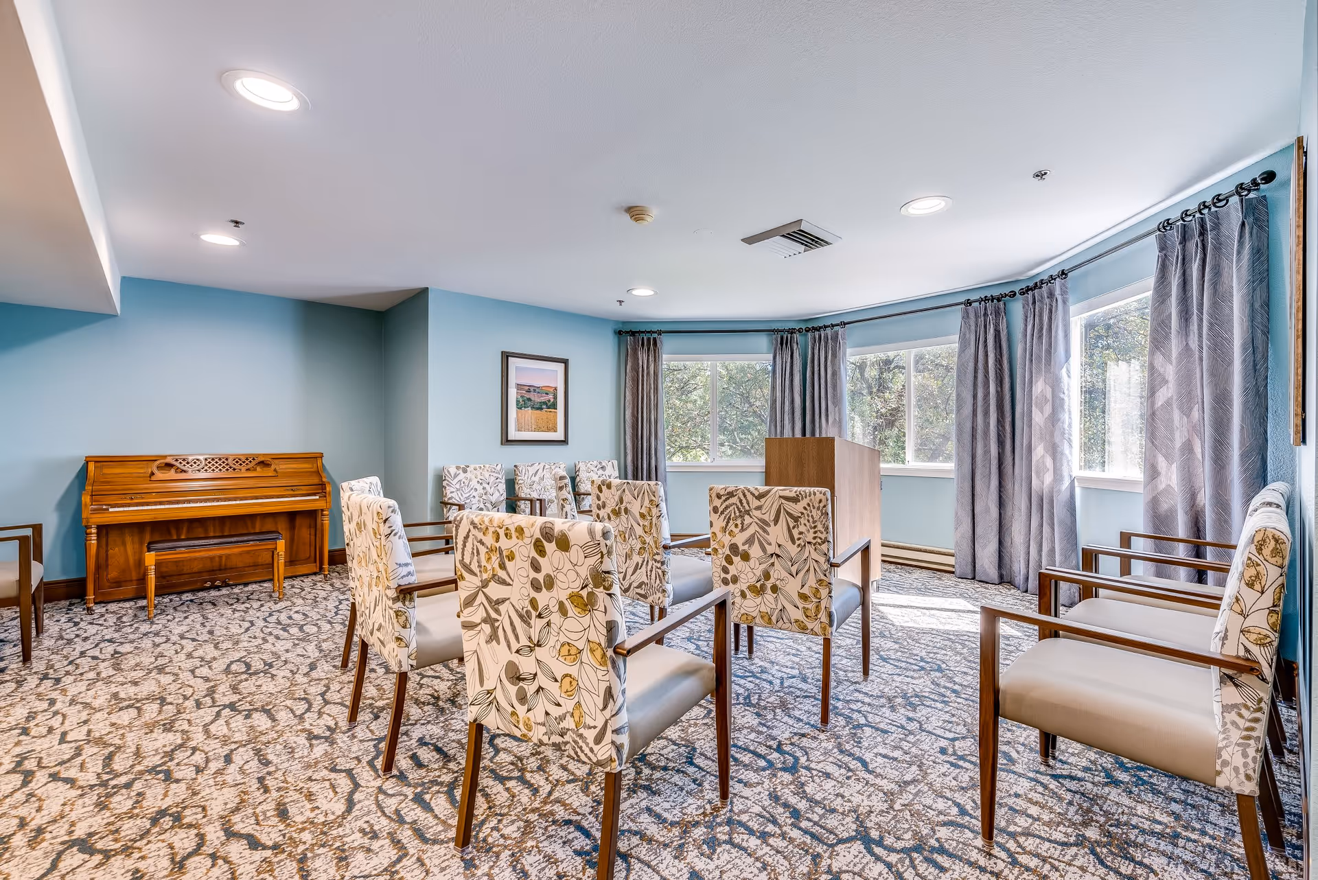 A bright room with patterned carpet and light blue walls, featuring several upholstered chairs arranged in a semi-circle facing a wooden podium. A wooden piano with a bench is against one wall, and large windows with gray curtains allow natural light to fill the space.