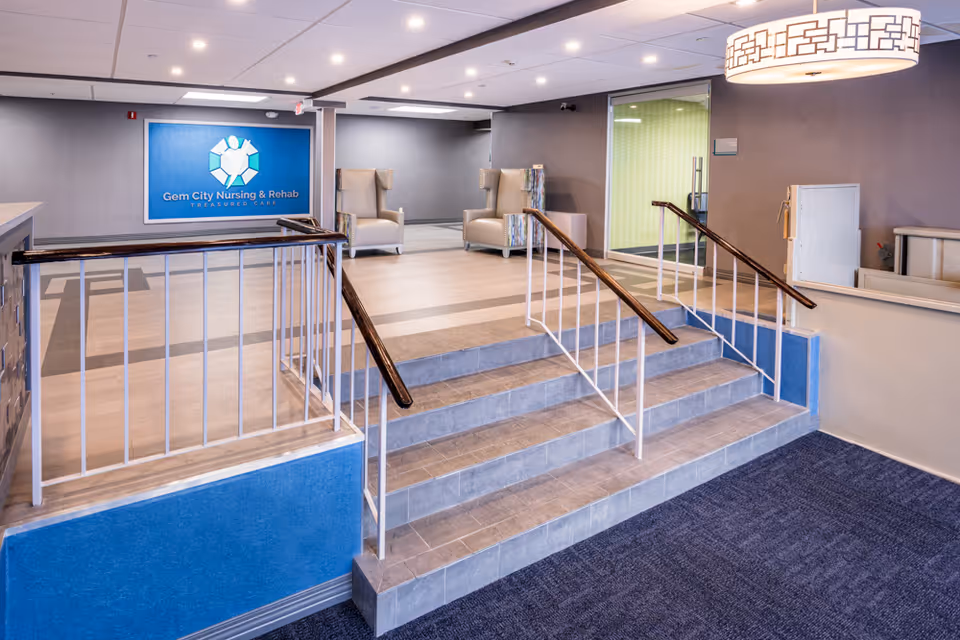Interior lobby of Gem City Nursing & Rehab with a short tiled staircase and handrails leading up to a seating area and a blue facility sign on the back wall.