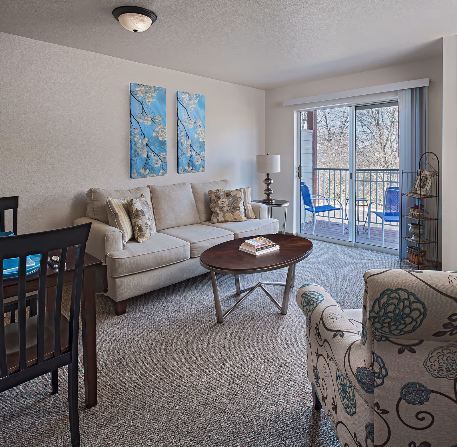 A cozy living room with a beige sofa adorned with patterned pillows, a wooden oval coffee table with books on it, a floral-patterned armchair, and a side table with a lamp. Two blue floral paintings hang on the wall above the sofa. Sliding glass doors lead to a balcony with two blue chairs and a small table, overlooking trees outside.