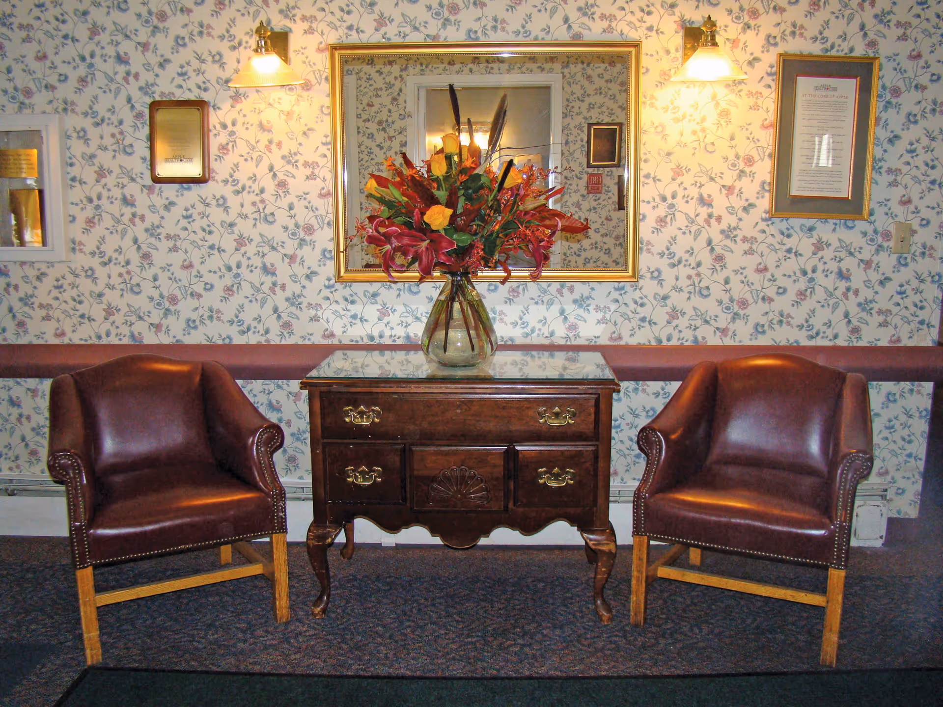 Two brown leather armchairs flank a wooden side table with a glass top, which holds a vase of colorful flowers. The background features floral wallpaper, a large framed mirror, two wall-mounted lights, and framed documents on the wall.