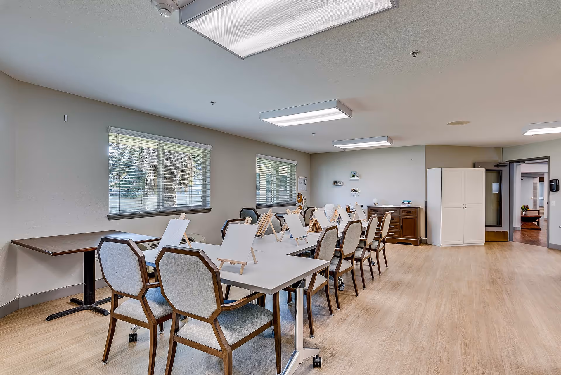 A bright activity room in a senior living facility with a long white table surrounded by chairs. Each chair has a small easel with a blank canvas set up for painting. The room has large windows with blinds, light wood flooring, and fluorescent ceiling lights. In the background, there is a wooden cabinet and a white storage cabinet near an open door.