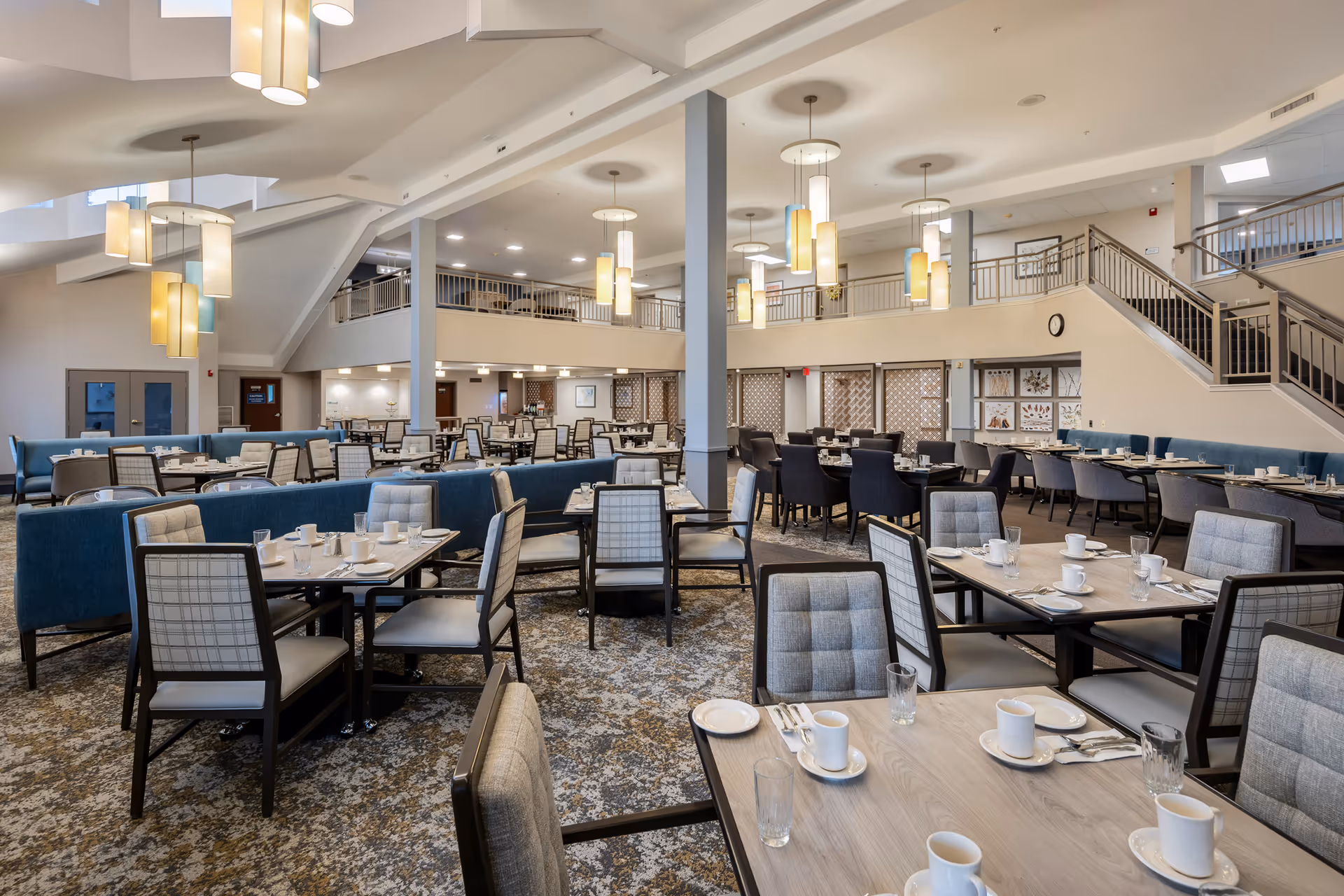 Spacious dining room with multiple tables and chairs arranged neatly, set with white cups, plates, and silverware. The room features modern pendant lighting, a patterned carpet, and a staircase leading to an upper level with railings. The walls have decorative panels and the overall atmosphere is bright and welcoming.