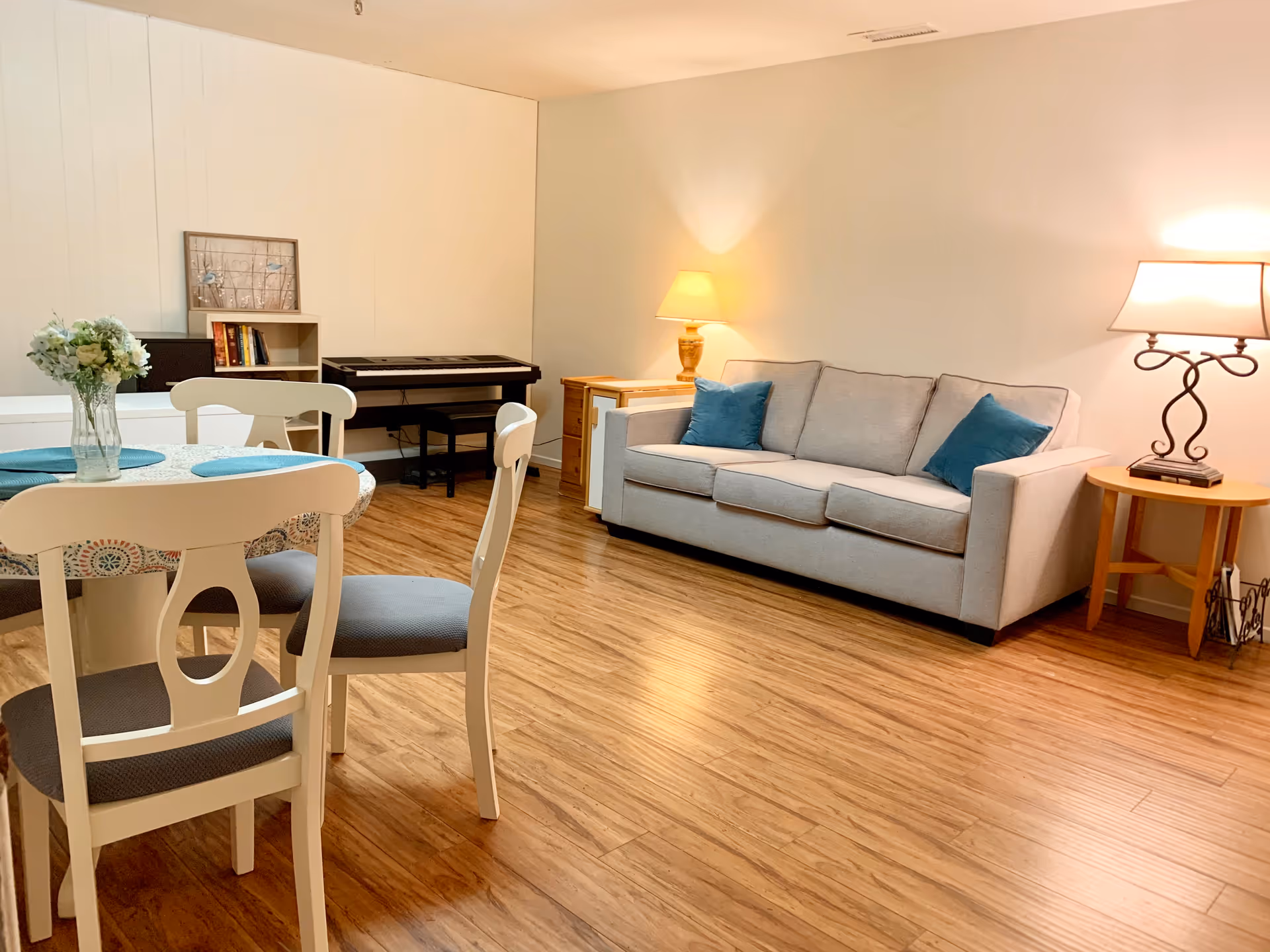 A cozy living room area with a light gray sofa adorned with two blue throw pillows, two side tables each with a lamp, a round dining table with four white chairs, and a small bookshelf with a keyboard piano on top. The floor is wooden and the walls are light-colored.