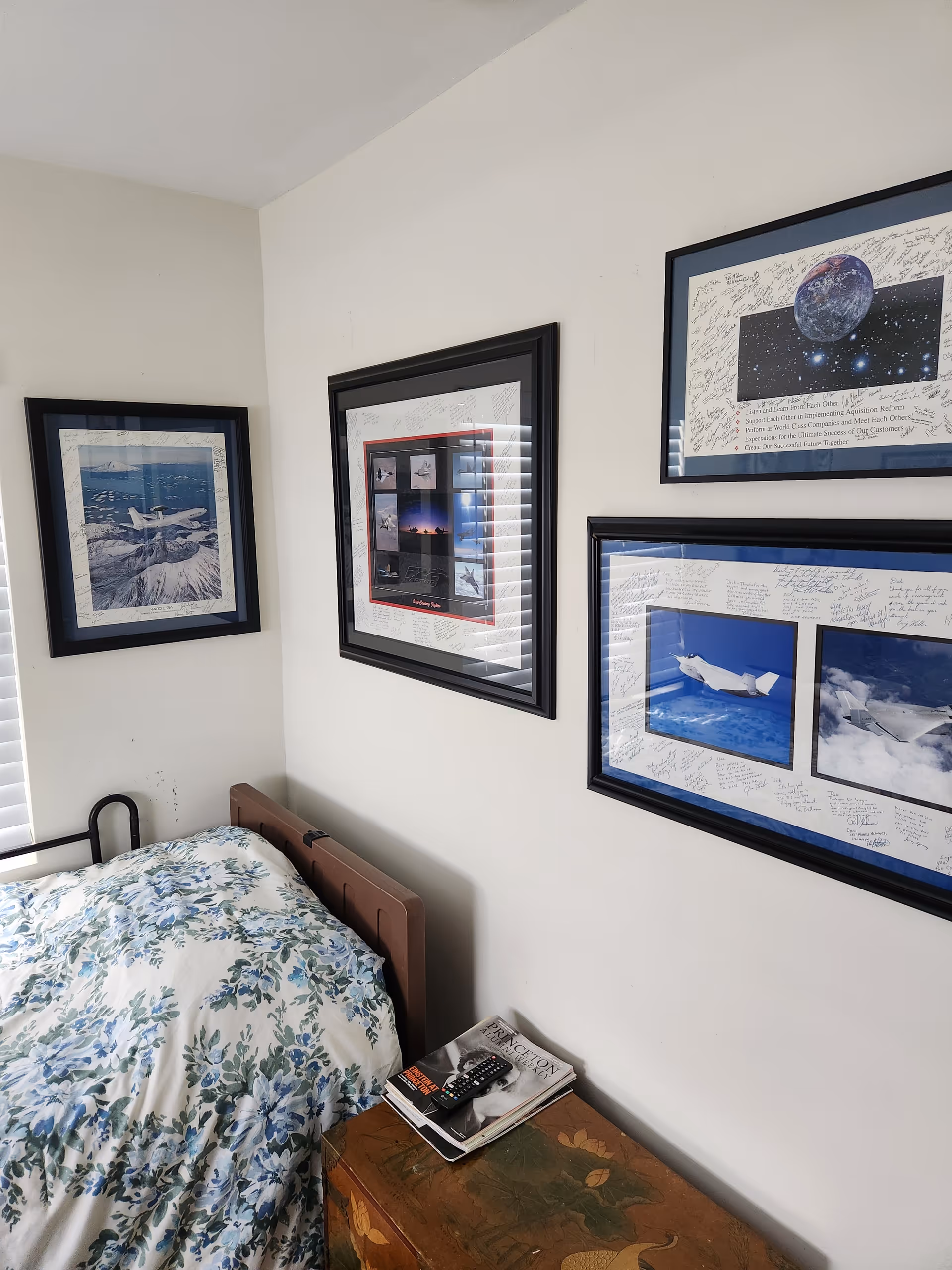A corner of a bedroom with a bed covered in a floral patterned comforter. Above the bed, there are four framed pictures on the walls, featuring images of airplanes and space. A small wooden chest next to the bed holds a remote control and some magazines.