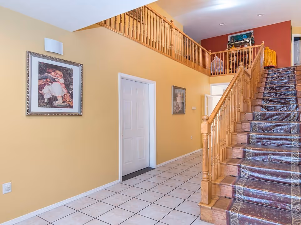 Interior view of a hallway with yellow walls and white tiled floor. A wooden staircase with a patterned carpet runner leads to an upper floor with wooden railings. Two framed pictures hang on the walls, and there is a white door along the hallway.