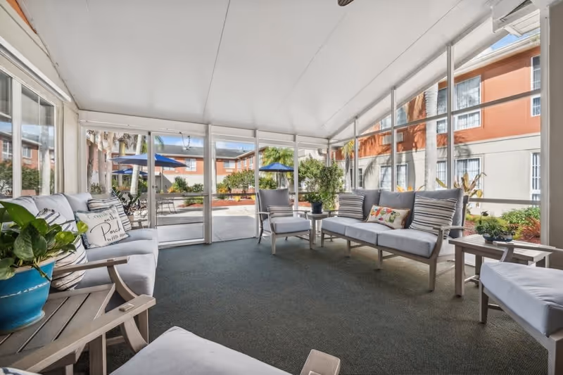A bright and airy enclosed patio area with cushioned seating including sofas and chairs arranged around small tables. The space has large windows providing a view of an outdoor courtyard with umbrellas, plants, and a building exterior in the background.