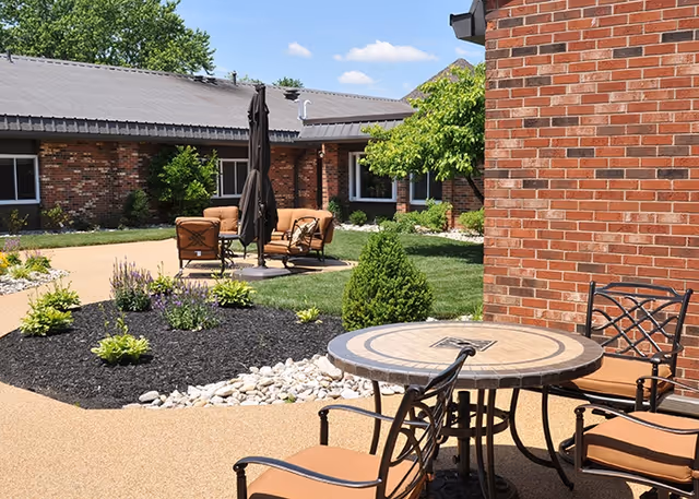 Outdoor patio area with a round table and four chairs in the foreground, and a seating area with cushioned chairs and a closed umbrella in the background. The area is surrounded by a garden with small bushes, plants, and decorative white rocks, adjacent to a brick building under a clear blue sky.