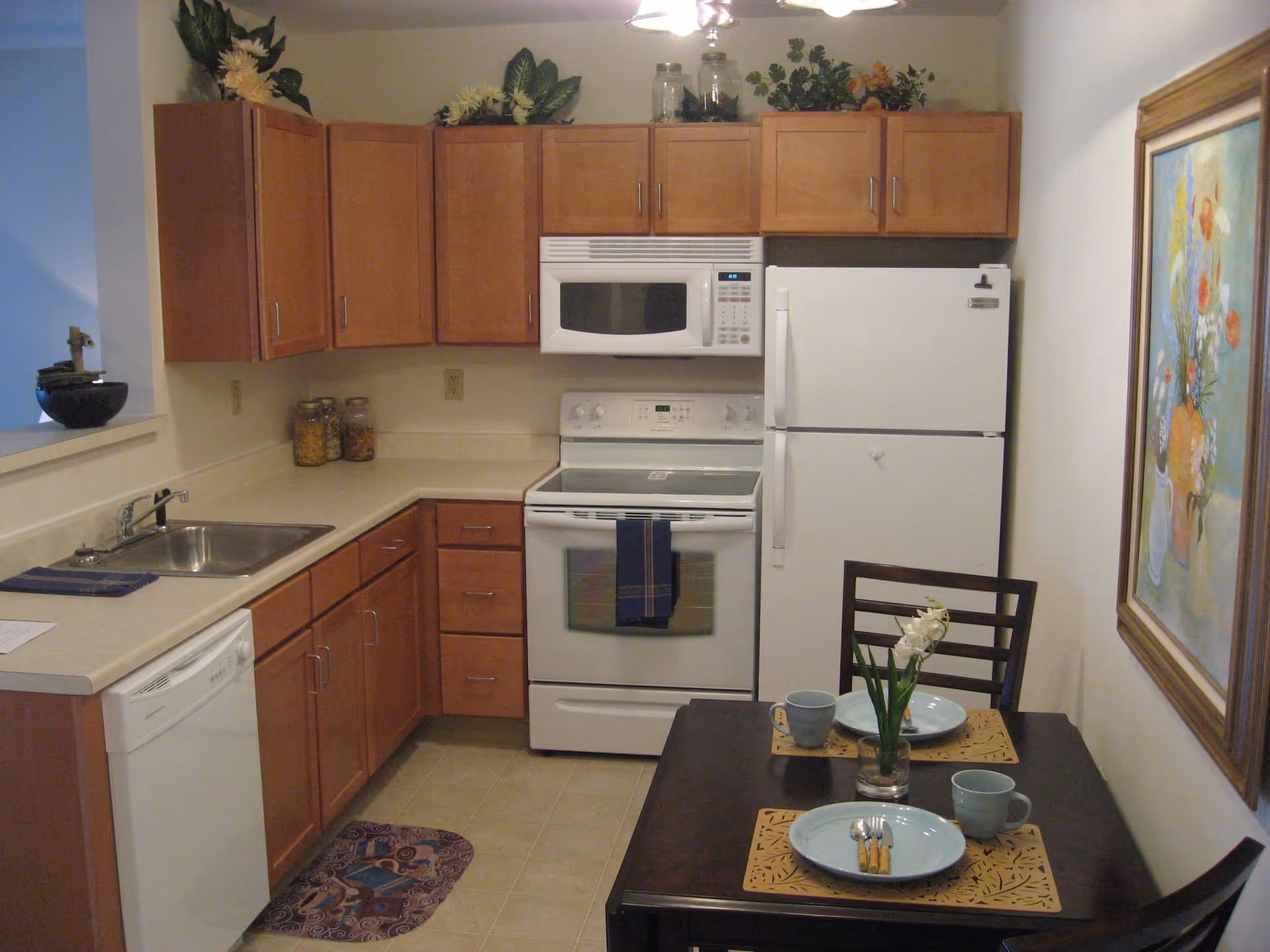 A compact kitchen with wooden cabinets, a white refrigerator, stove, microwave, and dishwasher. There is a small dining table set for two with blue plates, mugs, and a small flower vase. A colorful painting of flowers hangs on the wall beside the table.
